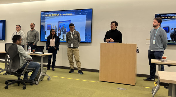 5 postdoctoral speakers standing near a podium answer questions from a live audience in front of computer monitors.