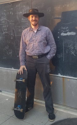 Donald Fershcweiler stands in front of a chalkboard holding a wooden skateboard.