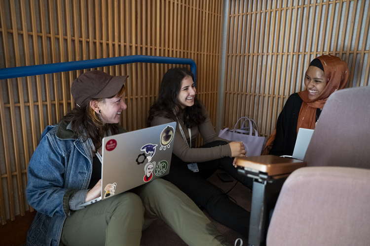 Three women of different ethnicities and cultures joyfully discuss computing over their laptops.
