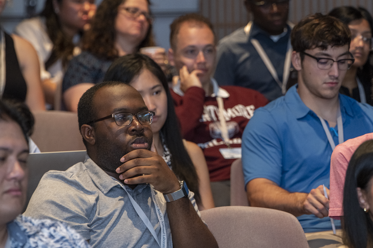 A diverse group of men and women of different ethnicities thoughtfully watch a presentation from their auditorium seats.