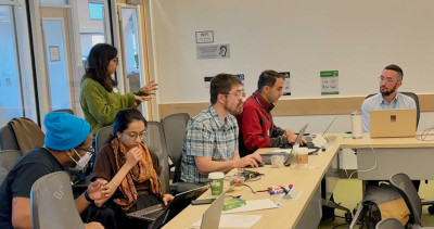 A group of people sitting around a conference table with laptops and notes, engaged in discussion. One person stands while addressing the group.