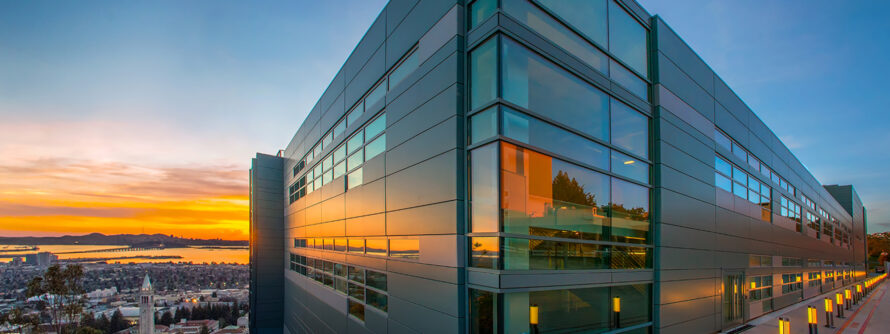 A modern building with glass panels stands on a hill above UC Berkeley, reflecting sunset sky colors in the glass.