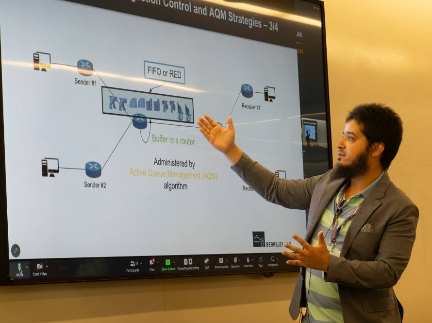 A young man with a beard and suit jacket gestures to a scientific diagram on a large monitor screen.