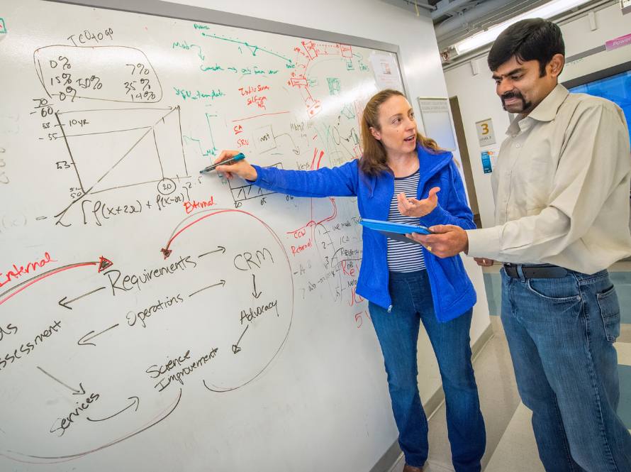 A woman points to a diagram on a white board while explaining to a man holding an iPad.