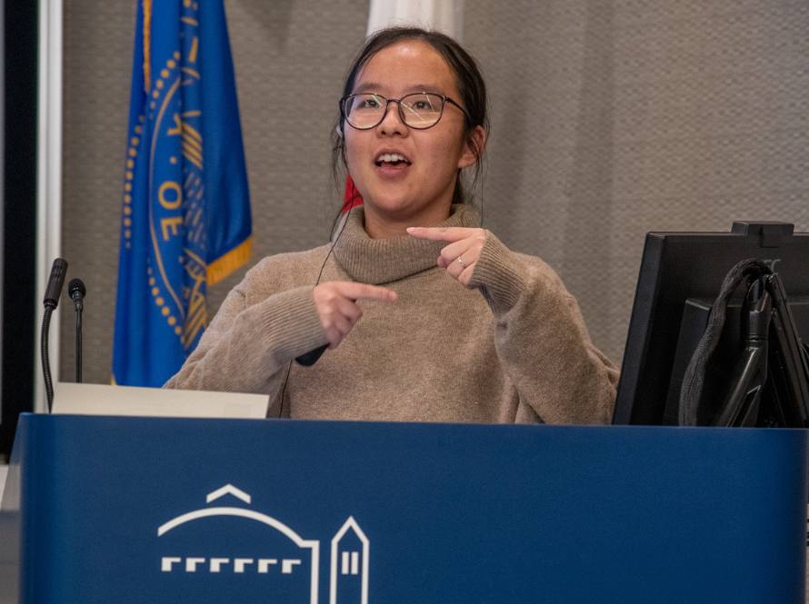 A young woman wearing glasses and a beige sweater speaks at a podium during the 2023 Postdoc Symposium event.