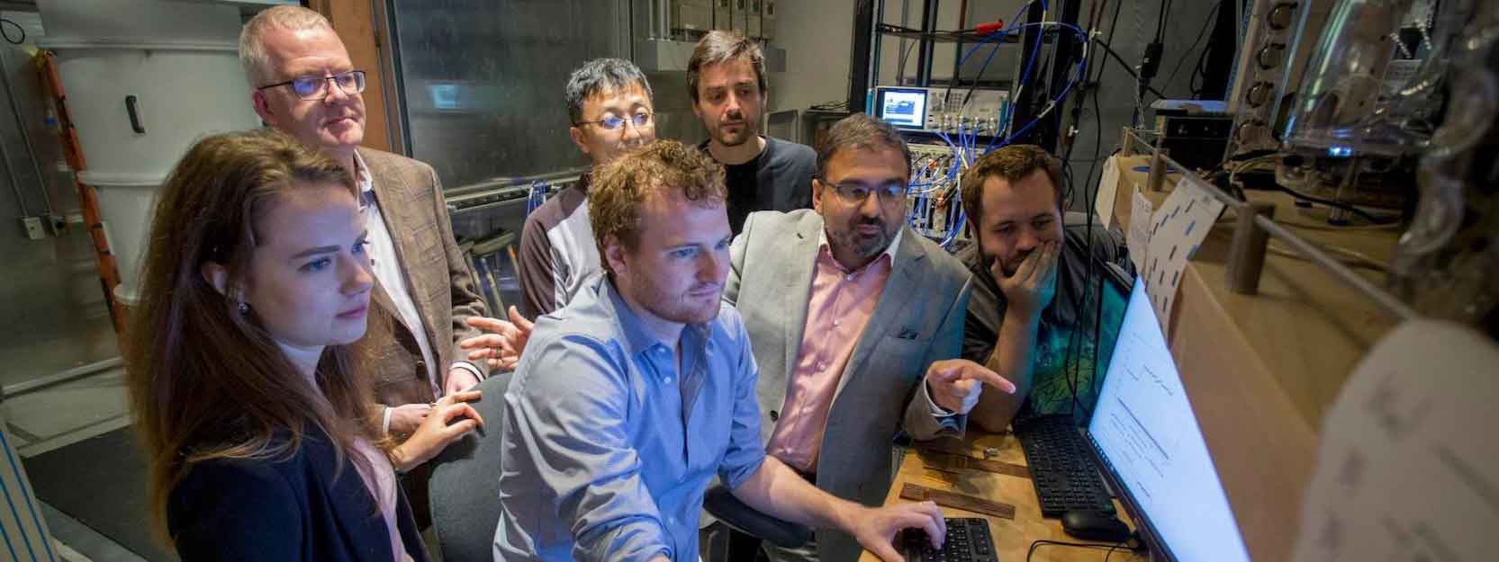 A diverse group of seven scientists look at a shared computer screen at a quantum research facility.