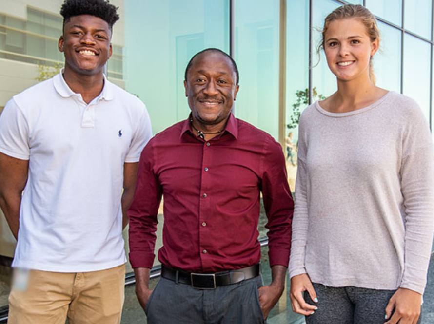 Two young adults, a black man and a caucasian woman, stand with an older black man in front of a lab building.