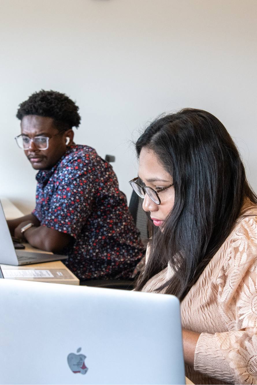 A young black man wearing glasses and a young woman of color wearing glasses sit together at workstations working on silver laptops at Shyh Wang Hall.