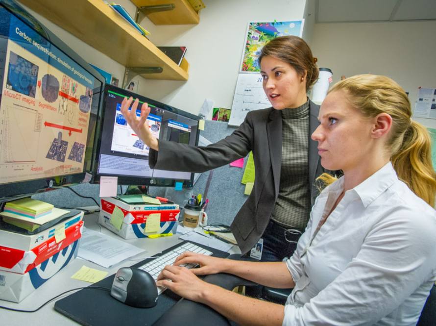 Two fair-skinned young women discuss carbon sequestration over a computer monitor.