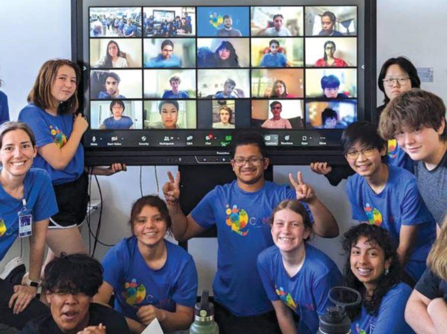 Group of young people participating in QCamp 2024 clustered around a large display of their fellow campers who are remote.