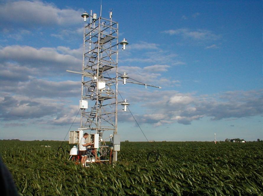 Photo of a researcher standing at the base of the AmeriFlux experiment tower, which stands alone in a field of green grass against a blue sky with clouds.