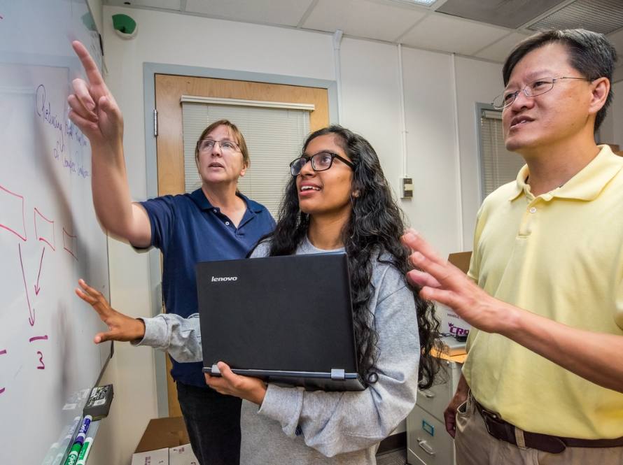 Two senior LBNL scientists, a man and a woman, discuss scientific findings on a white board with a young woman holding a laptop.