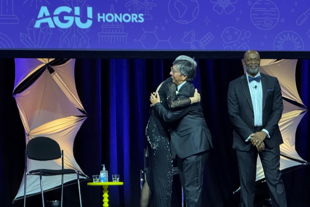 People on an awards stage in formal wear hugging after one of them is honored for their achievement. Behind then is a purple banner that says AGU Honors