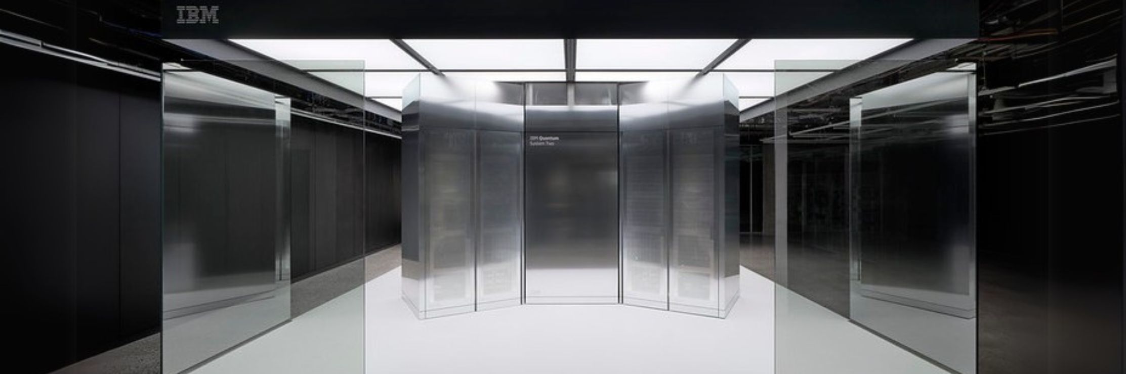 A sleek, futuristic IBM Quantum System Two computer is showcased in a dimly lit, high-tech environment. The system's reflective glass panels and minimalist design give it a cutting-edge, modern appearance. The IBM logo is visible at the top, emphasizing the branding of this advanced quantum computing system.