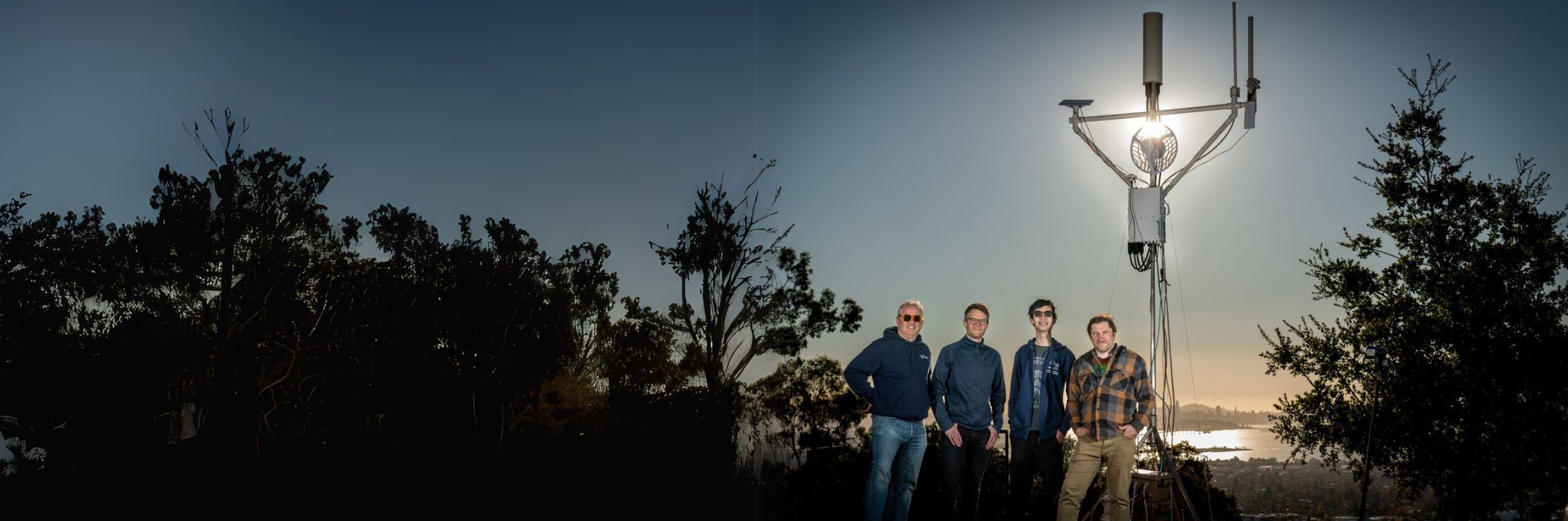 Four researchers stand in front of a wireless communication tower at sunset, overlooking a cityscape with a body of water in the background. The sun shines directly behind the tower, creating a dramatic silhouette effect. The group is dressed casually, with some wearing jackets and sunglasses.