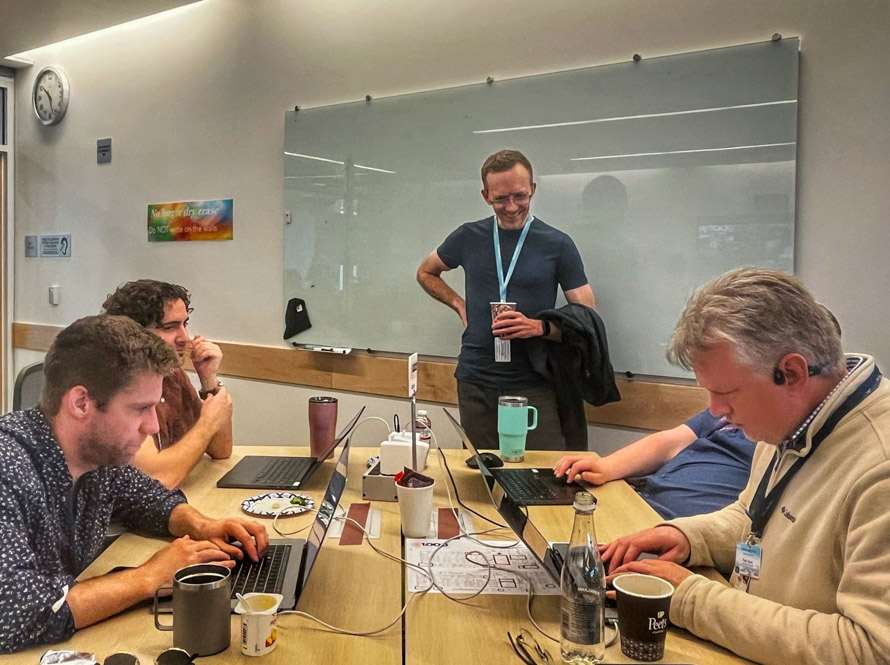 5 people gathered around laptops and a white board participate in the AI Jam event.
