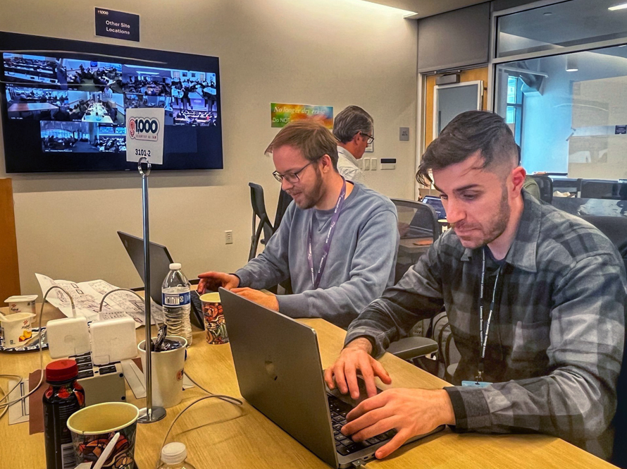 Two people enter prompts on their laptops in front of a Zoom screen displaying other participants across the United States during the AI Jam event.