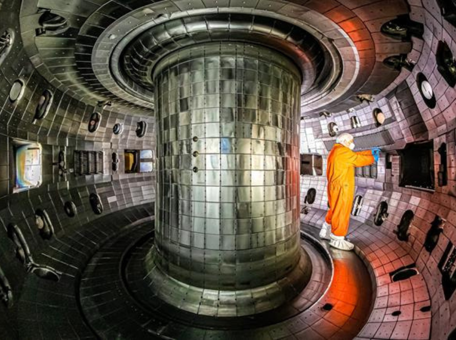 A person dressed in an orange protective suit and white boots is standing inside a large metallic chamber, which appears to be a fusion reactor. The chamber is cylindrical with a central column and is lined with numerous panels and components.