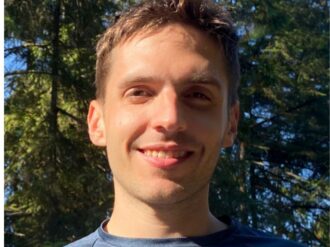 A person standing outdoors in front of tall evergreen trees, with sunlight filtering through the branches.