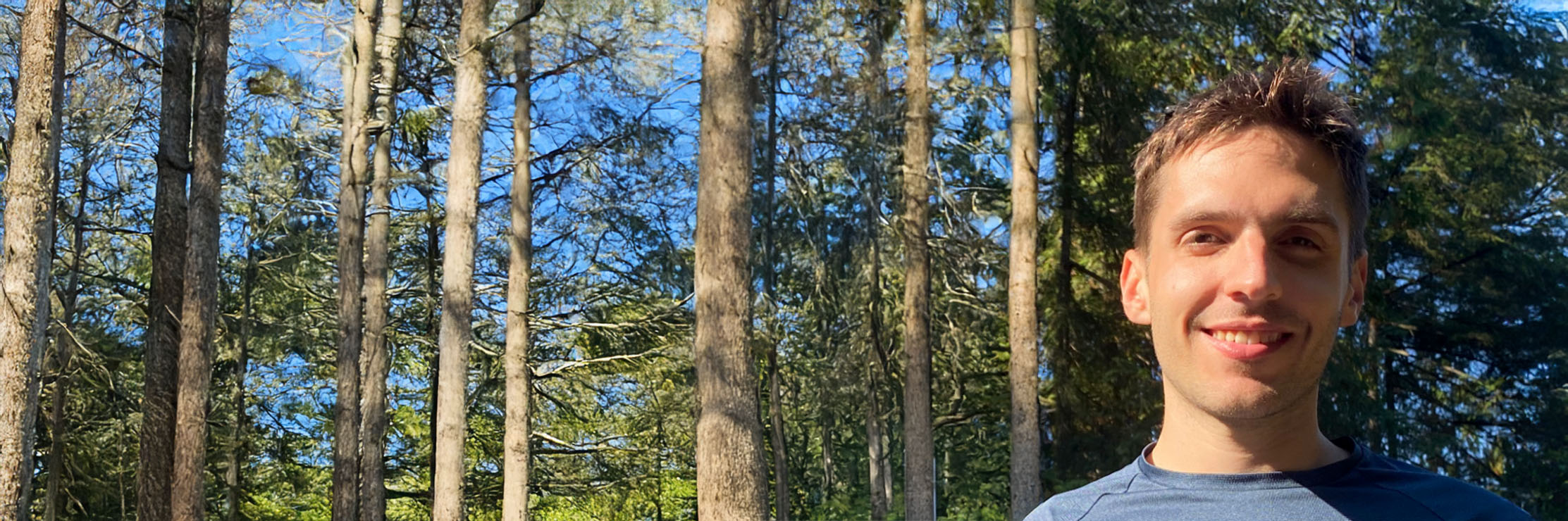A person standing outdoors in front of tall green trees, wearing a dark crew-neck shirt in natural sunlight.