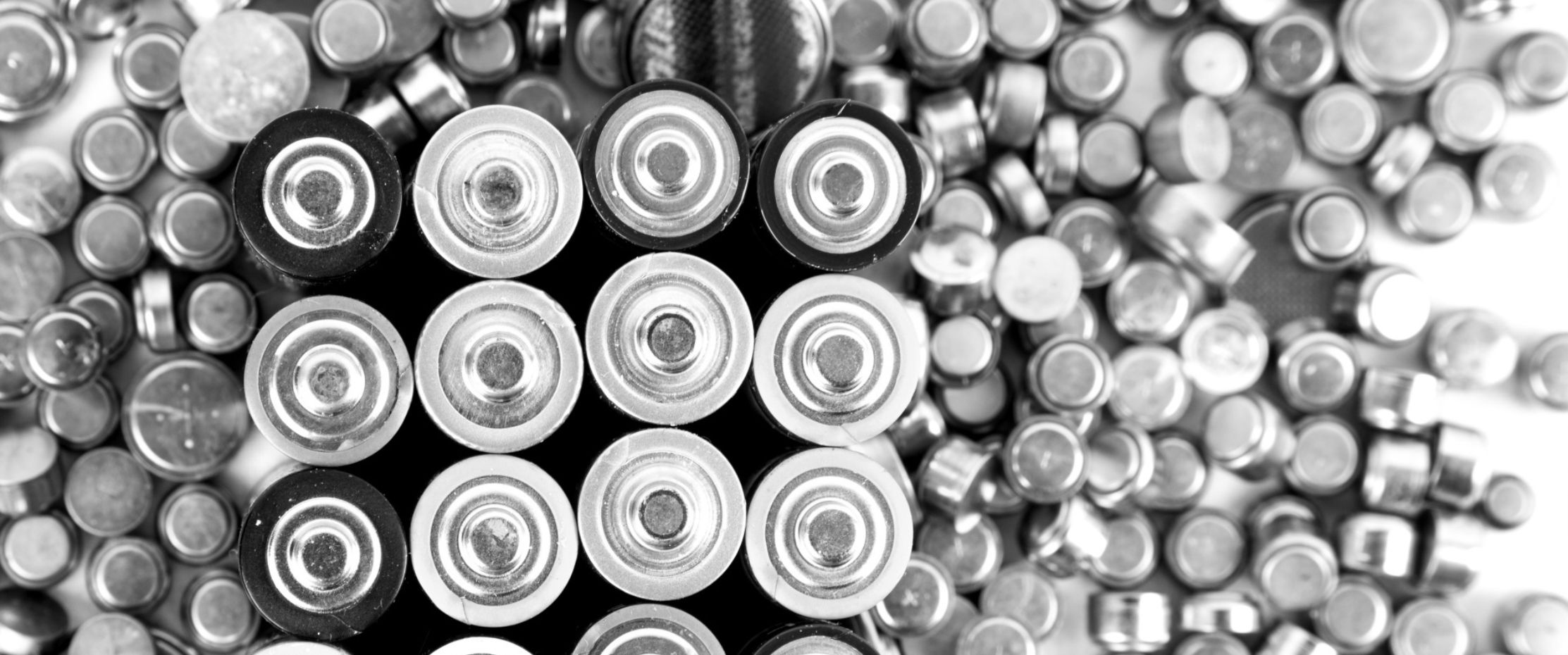 Close-up, black and white image showing the tops of various cylindrical batteries, including AA and button cell types, arranged in a cluster.
