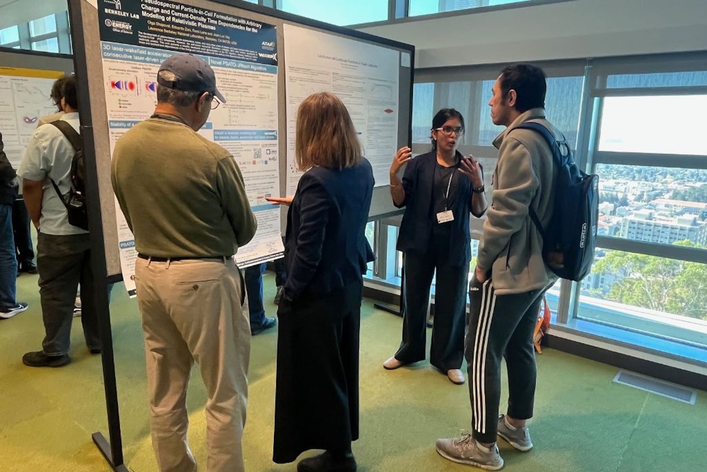 Three people stand in front of a large scientific poster, listening and engaging with a presenter who is explaining her research. The poster session takes place in a bright room with floor-to-ceiling windows, providing a view of city buildings and trees outside. Other attendees and research posters are visible in the background.