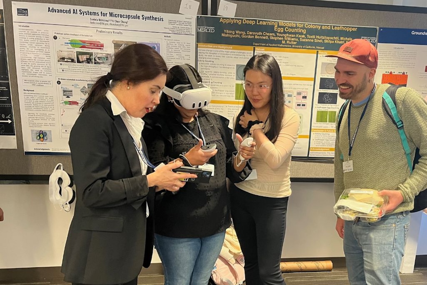 Four people stand in front of scientific research posters at a conference. One person is wearing a VR headset and holding controllers, while another is interacting with them and holding a phone. The others are engaged in conversation; one is holding a sandwich and wearing a backpack. The posters behind them discuss Daniela Ushizima, a Berkeley Lab scientist, stands at the far left demonstrating AI systems for microcapsule synthesis to three conference attendees. One attendee wears a VR headset and holds controllers while others observe and discuss. Behind them, scientific research posters are displayed on a bulletin board.