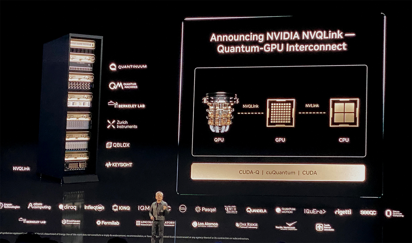 A presenter speaks on a dark stage next to a large server rack and a projected screen announcing NVIDIA NVQlink, a quantum-GPU interconnect. The visual shows partner organizations and a diagram connecting a quantum processing unit (QPU), graphics processing unit (GPU), and central processing unit (CPU).