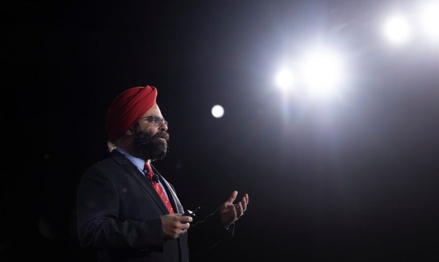 A person wearing a red turban, a suit, and a polka-dot tie stands on stage giving a presentation, gesturing with one hand while holding a remote control in the other, with a microphone clipped to the jacket and a spotlight shining behind.