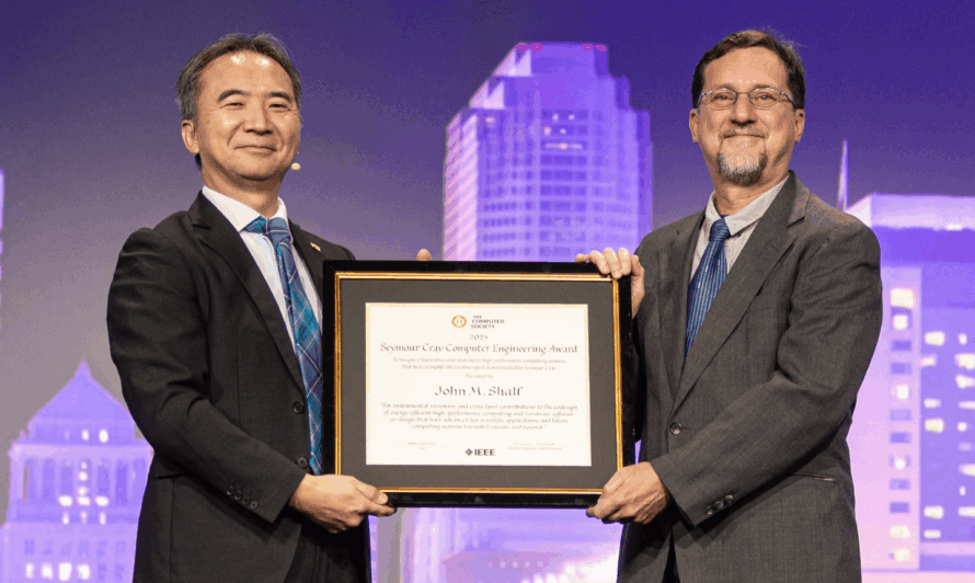 Two people in suits stand on a stage, with one handing an award plaque to the other. A large screen in the background displays the recipient's name and job title along with the name of their institution.