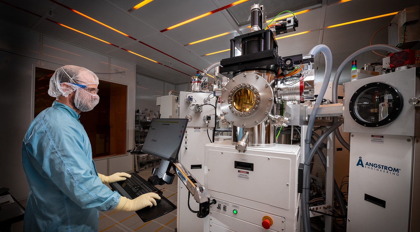 Individual in a blue cleanroom suit and yellow gloves operating a computer in front of an advanced vacuum deposition machine inside a clean laboratory.