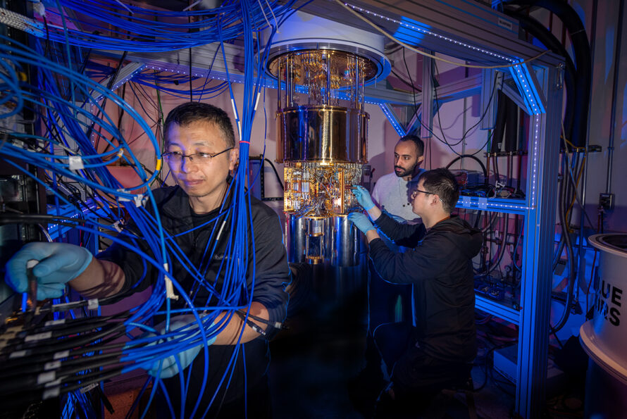 Several individuals wearing gloves working around a large, cylindrical metallic quantum computing dilution refrigerator with numerous bright blue cables and illuminated hardware in a high-tech lab environment.