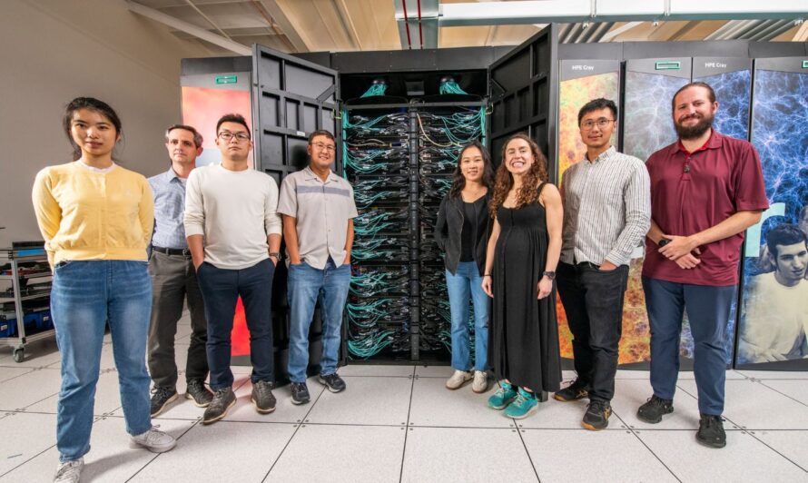 A group of people standing in front of a large supercomputer system, with visible racks of cables and colorful panels featuring scientific imagery in the background, inside a modern research facility.