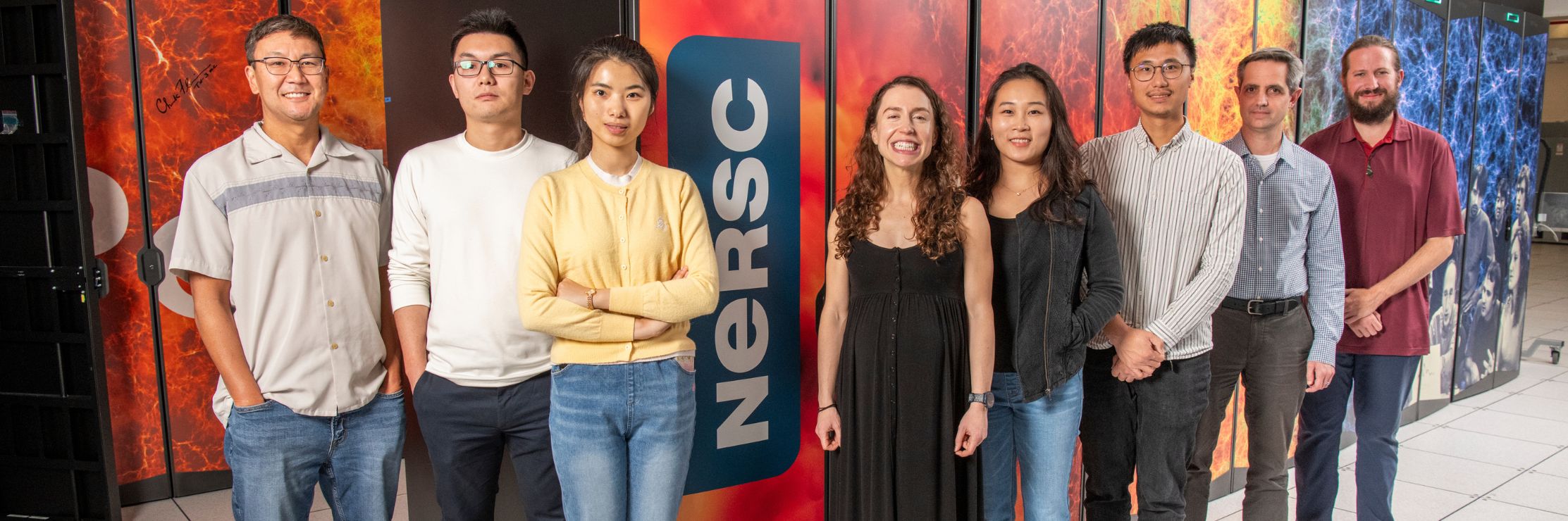 A group of people standing in front of a large supercomputer system, with visible racks of cables and colorful panels featuring scientific imagery in the background, inside a modern research facility.