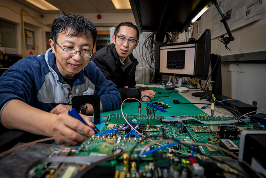 Two people work in a laboratory at a workstation covered with electronic circuit boards and cables, with waveform data displayed on monitors in the background.