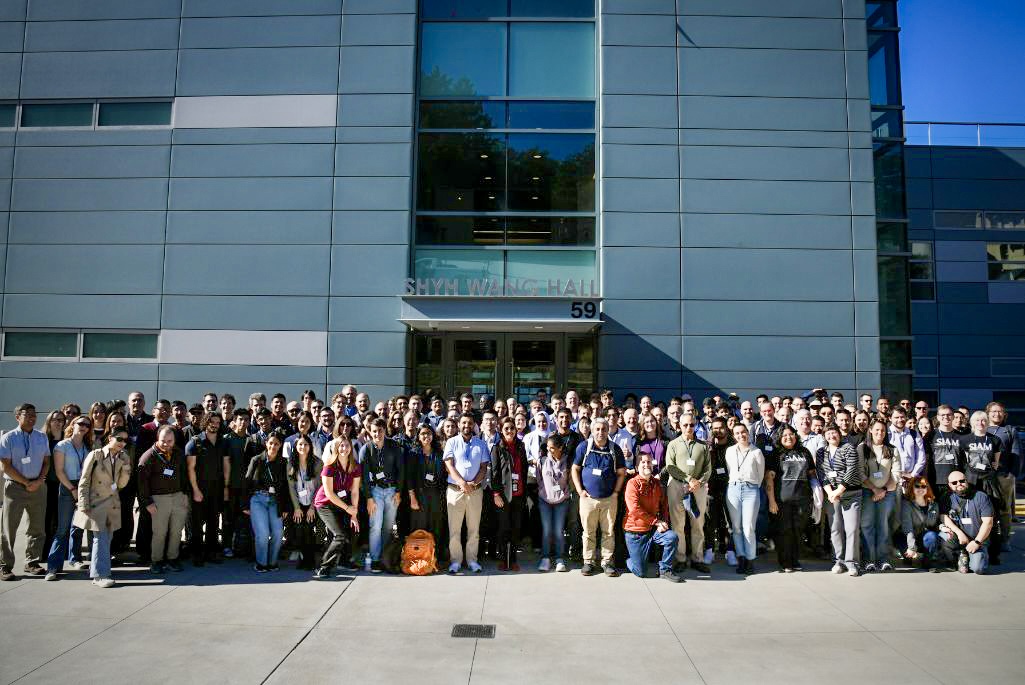 A large group of people pose together for a group photo in front of a modern building labeled "SHYH WANG HALL 59." The group is diverse, including men and women of various ages, and some people are wearing conference badges. The photo was taken on a sunny day, and the building features large windows and gray metal paneling.