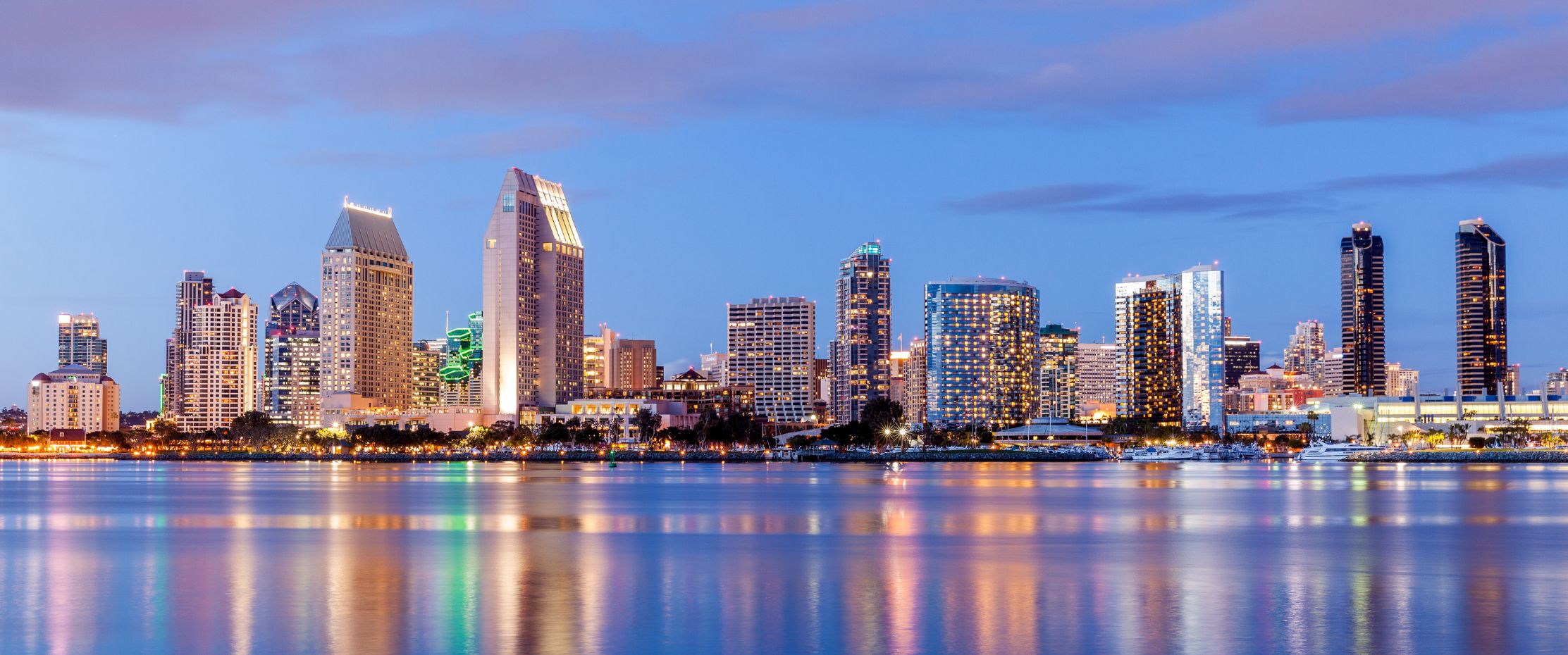 San Diego skyline at dusk, with tall buildings illuminated and reflections shimmering on the calm water in the foreground against a partly cloudy blue sky.