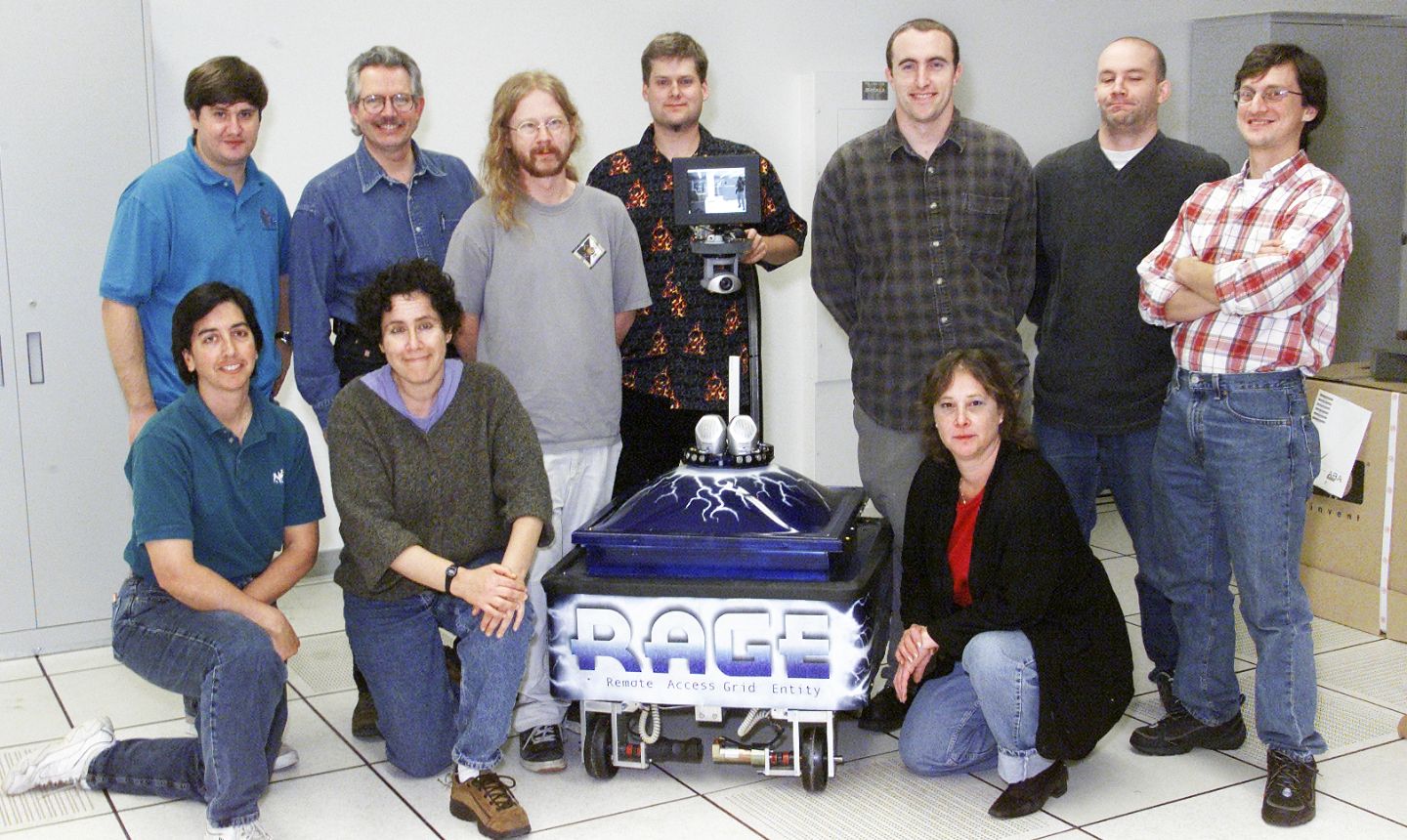 A group of people pose in a computer lab around the Remote Access Grid Entity (RAGE) robot, a blue mobile device with a mounted camera and display screen, designed and built by staff from Berkeley Lab's NERSC, Computational Research, and Information Technologies and Services divisions.