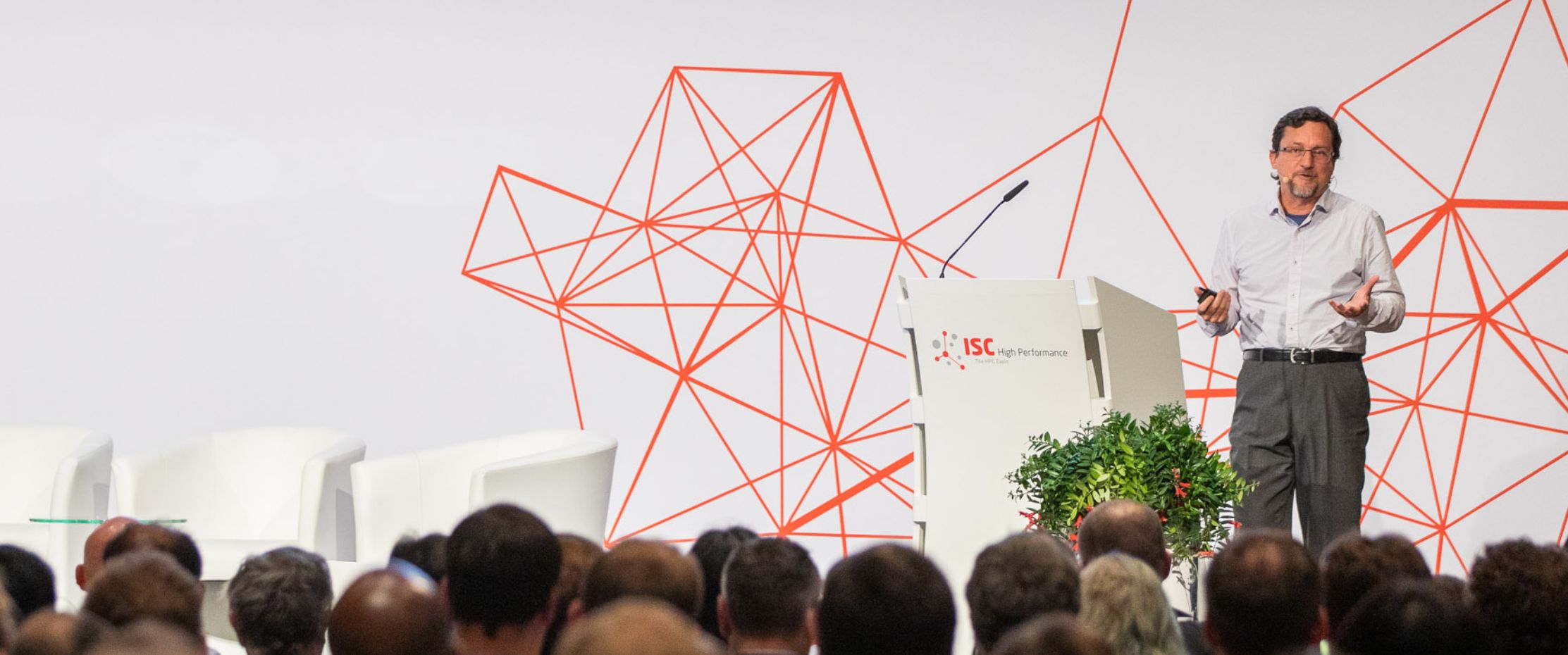 A person in a light-colored button-down shirt stands on stage wearing a headset microphone, with geometric red line art and a gradient-colored border visible in the background.