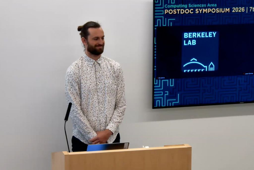 Alec Deckto stands behind a wooden podium at the Berkeley Lab Postdoc Symposium. He is wearing a white, patterned button-down shirt. To his right, a screen displays the event title and the Berkeley Lab logo on a blue digital background.