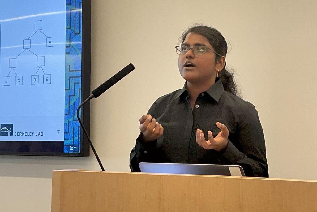 Durga Mandarapu, a woman with dark curly hair and glasses, speaks animatedly at a wooden podium. She is wearing a black collared shirt and gesturing with both hands. To her left, a presentation slide displays a tree diagram for a presentation at Berkeley Lab.