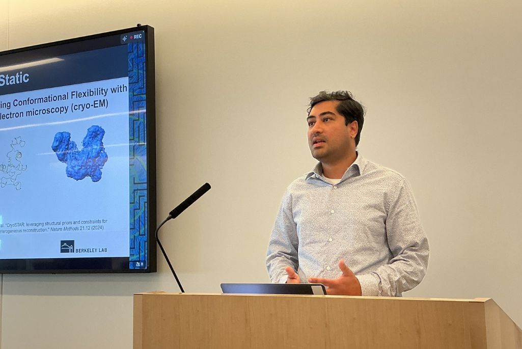 Nabin Giri gives a presentation, standing behind a wooden podium and gesturing with his hands. He has dark hair and is wearing a light-colored, subtly patterned shirt. To his left, a large screen displays a slide about using cryo-electron microscopy to map conformational flexibility, showing images of molecular models.