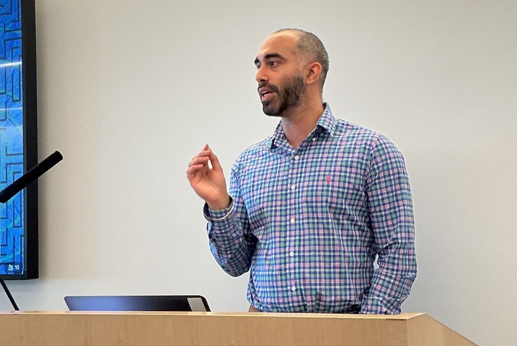 Navjot Singh, a man with a shaved head and a beard, gives a presentation from behind a wooden podium. He is wearing a blue and green plaid button-down shirt and is gesturing with his right hand to emphasize a point.