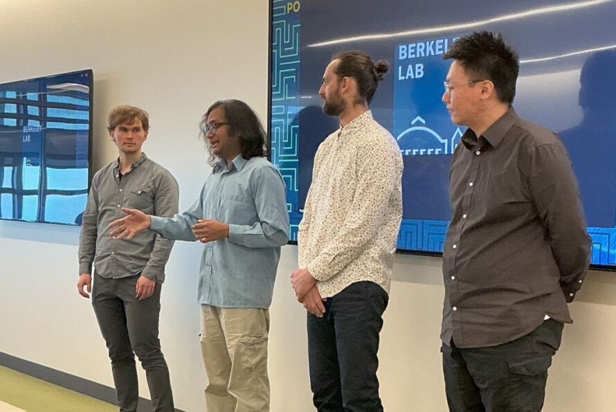 A group of five diverse male speakers stand in a line at a symposium, facing towards the left of the frame. They are dressed in business casual attire, including button-down shirts and slacks. A large monitor in the background displays an image of the group standing in the same formation.