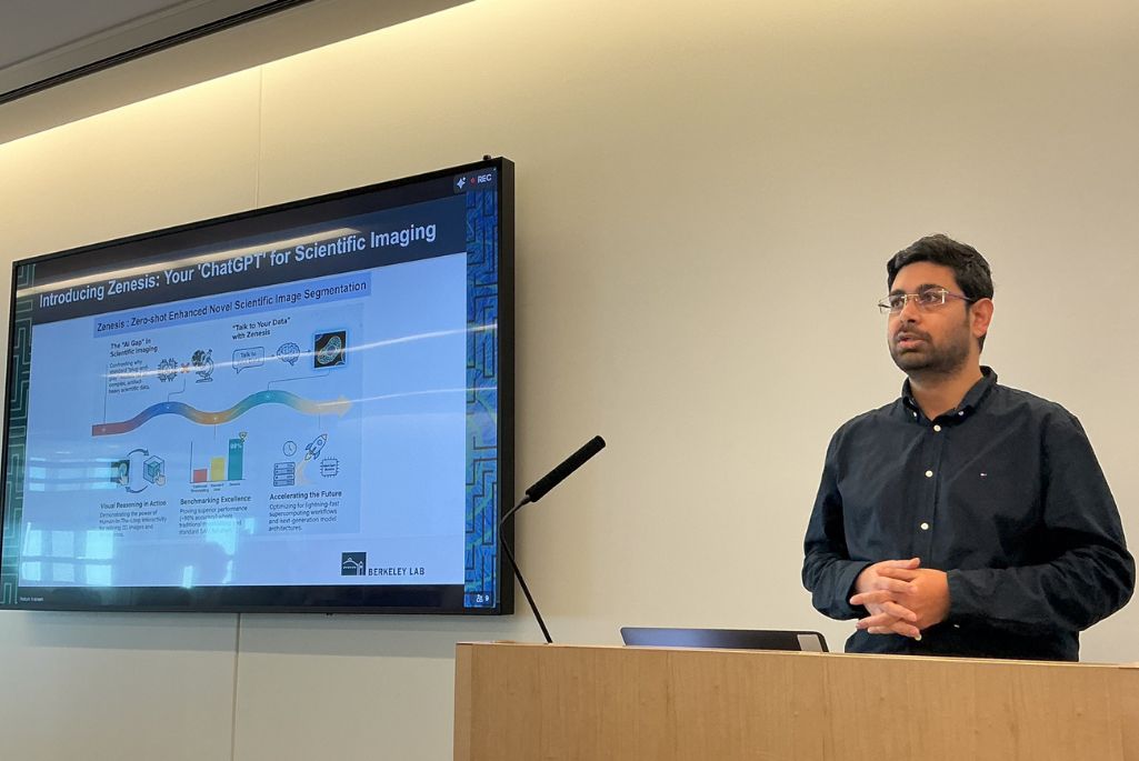  A man with short dark hair and glasses presents at a wooden lectern. He is wearing a navy blue shirt with the sleeves rolled up. Beside him, a presentation slide shows a colorful infographic with the title "Introducing Zenesis: Zero-shot Enhanced Novel Scientific Image Segmentation" and the Berkeley Lab logo.



