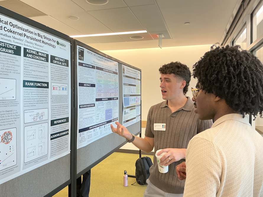 Two summer interns discuss a large scientific research poster.