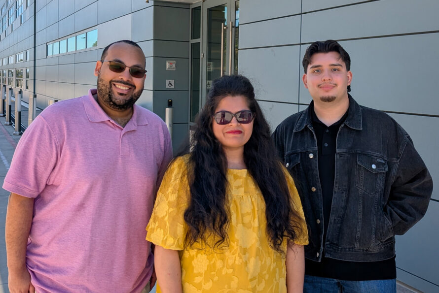 Three people stand smiling for a photo outside the entrance of a modern, grey-paneled building labeled "Shyh Wang Hall". From left to right: a man wearing sunglasses and a pink polo shirt, a woman wearing sunglasses and a bright yellow patterned top, and a young man wearing a black denim jacket over a black shirt with blue jeans. They are standing on a wide concrete walkway on a bright, sunny day.
