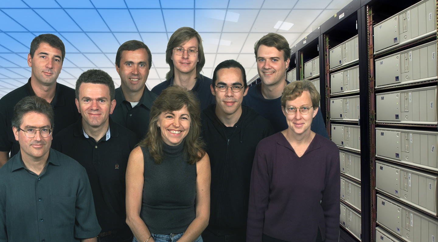 A group portrait of nine people casually dressed and smiling at the camera. They are posed together against a backdrop featuring a brightly lit server room filled with tall racks of computer equipment.