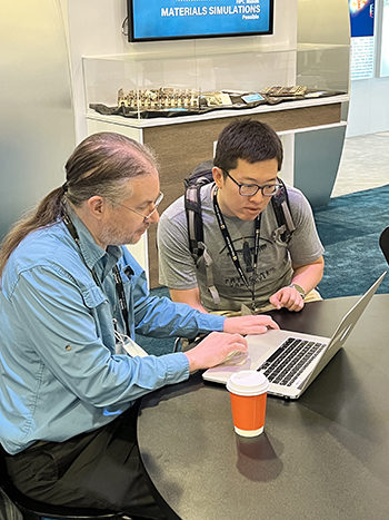 Two men wearing conference lanyards are sitting at a round black table looking at a silver laptop together. The man on the left in a blue shirt is typing, while the man on the right in a grey t-shirt watches the screen, with a coffee cup sitting nearby on the table.