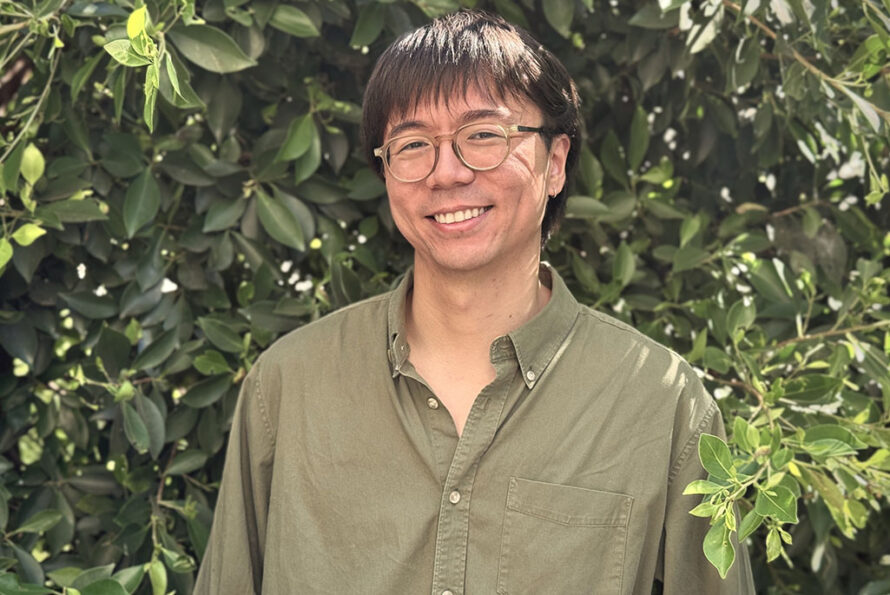 Samson Wang smiling warmly at the camera while standing outdoors in front of a backdrop of dense green leaves. They have short dark hair with bangs and are wearing round wire-rimmed glasses and an olive green button-down shirt.